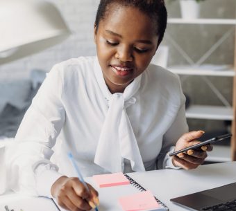 Skilled african-american graphic designer making sketch in notebook for new project using her mobile phone. Noting down important information. Modern lifestyle. Lady boss.